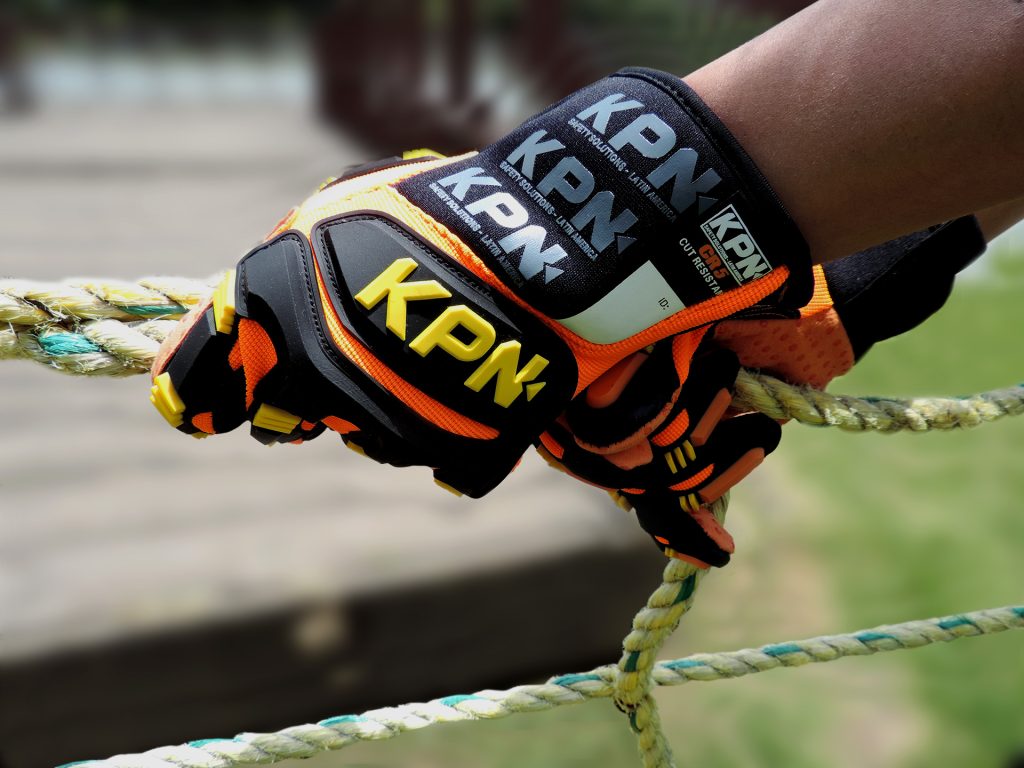 Close-up de uma mão usando luvas de proteção de alta visibilidade da marca KPN, nas cores laranja e preto, segurando firmemente uma corda grossa trançada. O fundo está levemente desfocado, mostrando um ambiente externo