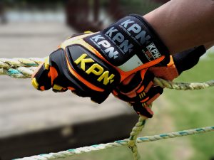 Luvas KPN, Close-up de uma mão usando uma luva de segurança de alta visibilidade da marca KPN, nas cores laranja e preto, segurando firmemente uma corda grossa trançada. O fundo está levemente desfocado, mostrando um ambiente externo