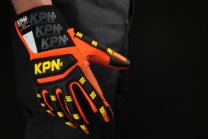 Luvas, mão vestindo uma luva de proteção técnica de alta visibilidade nas cores laranja e preto, posicionada de perfil contra um fundo escuro. O equipamento de proteção individual apresenta reforços emborrachados nos dedos e no dorso para mitigação de riscos de impacto e corte, fundamentais para a segurança em frentes de operação e manutenção na mineração. A imagem reforça a importância do monitoramento da integridade do trabalhador, evidenciando uma cultura de prevenção consolidada através de treinamento especializado e o uso de equipamentos de alta performance.