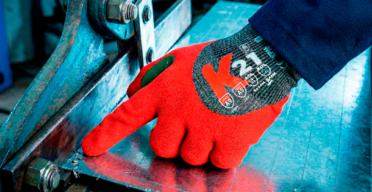 Mão com luva de segurança KPN vermelha manuseando peça metálica em ambiente industrial.